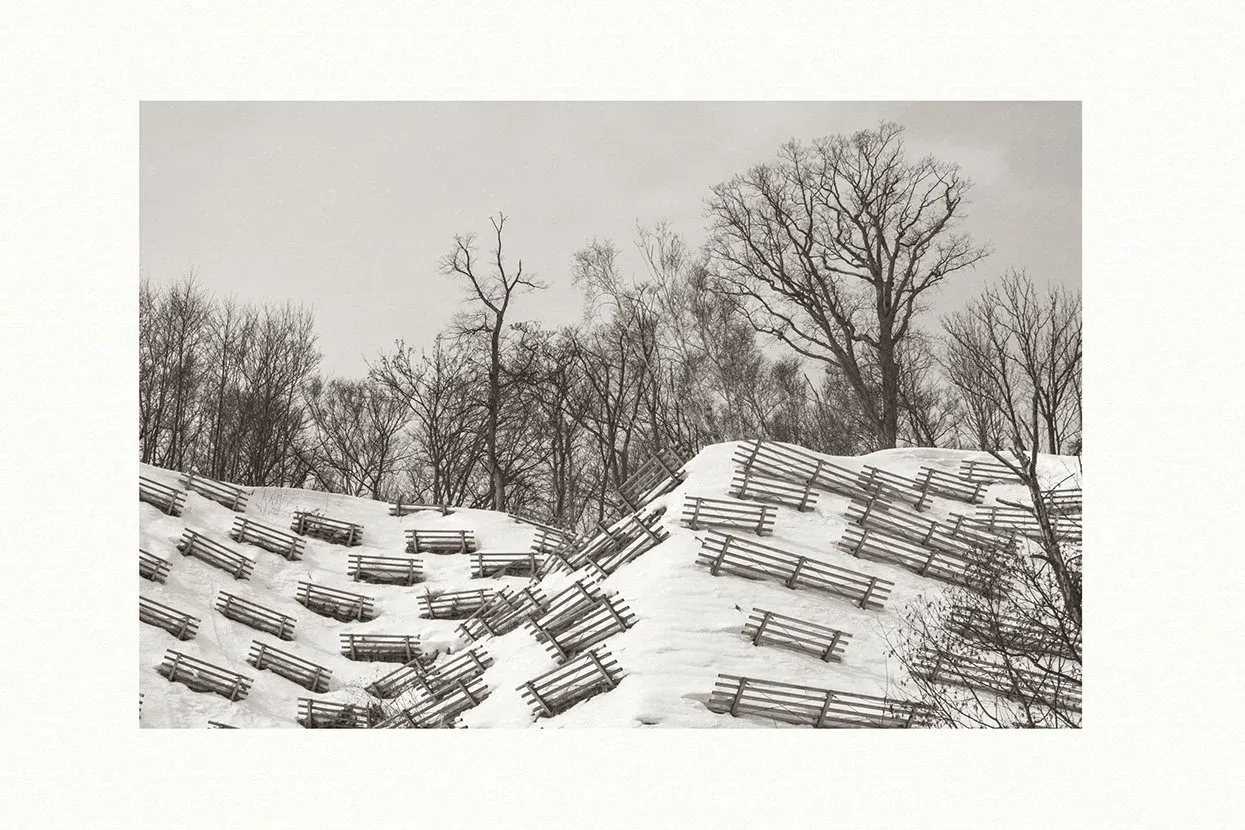 Warm-toned black and white print on Awagami Bizan: rows of wooden snow fences on a snowy hillside, bare trees under a grey sky